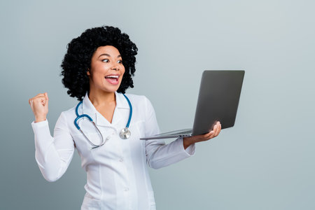 Cheerful female doctor celebrating achievement while holding laptop, standing against a gray background in a medical uniformの写真素材