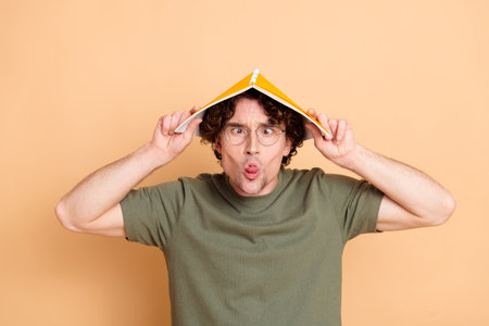 Playful young man making a funny expression by holding a book on his head and posing against a plain...