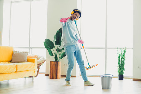 Young man enjoying household chores while mopping the living room in a cozy modern apartment interior on a sunny dayの写真素材