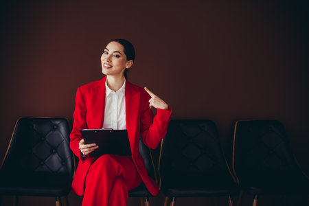 Beautiful Confident Young Businesswoman in Elegant Red Suit Sitting With Clipboard in Officeの写真素材