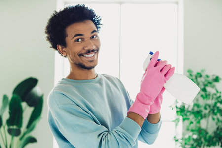 Young man wearing pink gloves holding a cleaning spray bottle while smiling in a bright modern living room interiorの写真素材