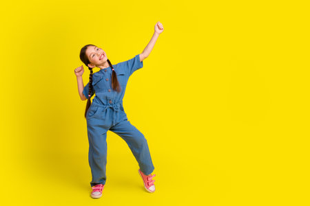 Adorable young girl posing with enthusiasm against a vibrant yellow background, dressed in stylish blue overalls and pink shoesの写真素材
