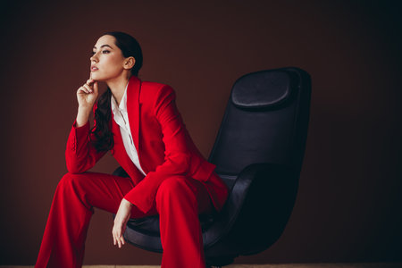 Beautiful Confident Businesswoman in Red Suit Sitting on Black Chair Against Brown Backgroundの写真素材