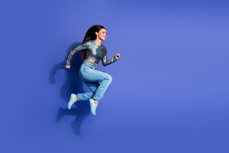 Vibrant young woman leaping with joy in a stylish outfit against a bright purple backdrop,...
