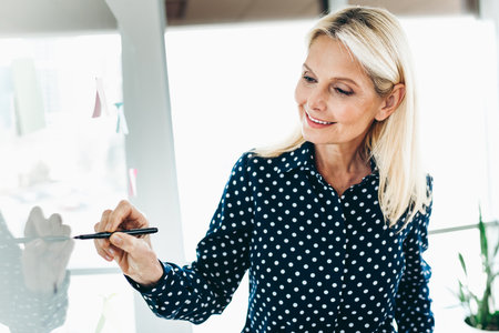 Professional businesswoman writing on a transparent board in a collaborative office setting during a daylight meetingの写真素材