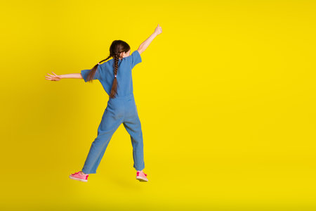 Energetic girl in trendy blue outfit jumping on yellow background with cheerful expression and playful gestureの写真素材