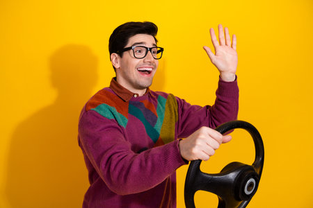 Young man with cheerful expression gestures while pretending to drive, wearing casual sweater styled...