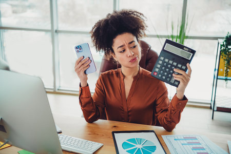Young professional businesswoman in office holding calculator and smartphone at desk analyzing data...
