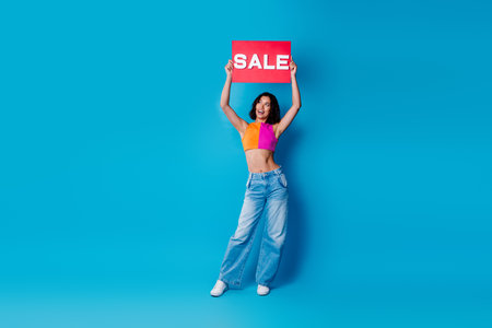 Cheerful stylish woman holding a red sale sign, posing against a vivid blue background, promoting a...