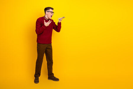 Young Male in Casual Sweater Expresses Frustration Using Smartphone Against Yellow Backgroundの写真素材