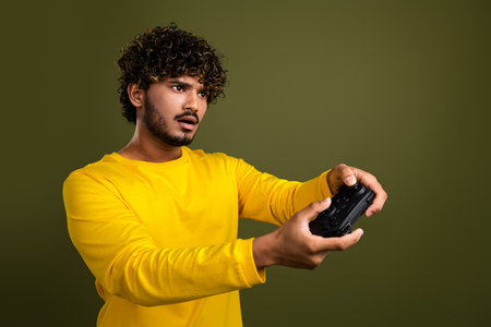 Young man concentrating while playing video games using a controller in a yellow shirt against a...