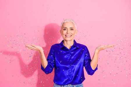 Confident mature woman in blue shirt celebrating with colorful confetti against a pink backgroundの写真素材