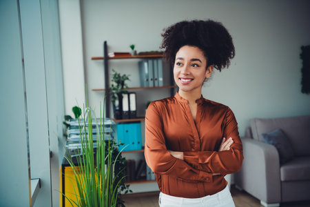 Confident businesswoman with curly black hair standing in a modern office with plants and bright daylightの写真素材
