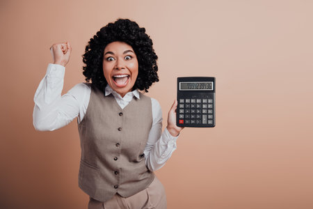 Excited Young Businesswoman Holding Calculator Celebrates Success Against Beige Backgroundの写真素材