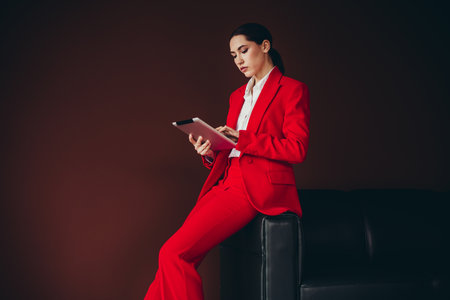 Confident Female Professional in Red Suit Using Digital Tablet Against Dark Backgroundの写真素材