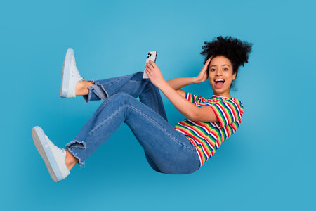 Cheerful young woman posing in mid-air with a smartphone on a bright turquoise background showcasing...