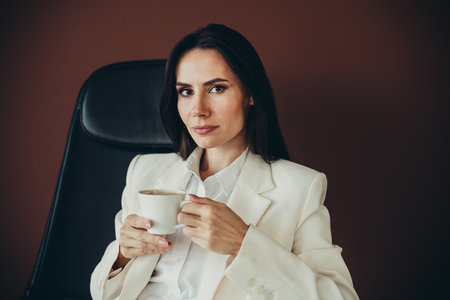 Confident businesswoman with a cup of coffee, stylishly dressed in a white suit, showcasing poise and elegance in a professional setting.の写真素材