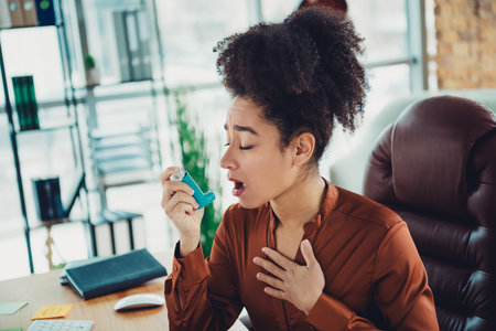 Young businesswoman using an inhaler in a modern office workspace with natural light, highlighting...