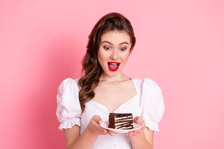 Pretty woman excitedly holding a chocolate cake slice on a plate with a pink background, expressing...
