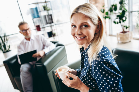Smiling mature businesswoman in stylish attire holding coffee in modern office interior with male...