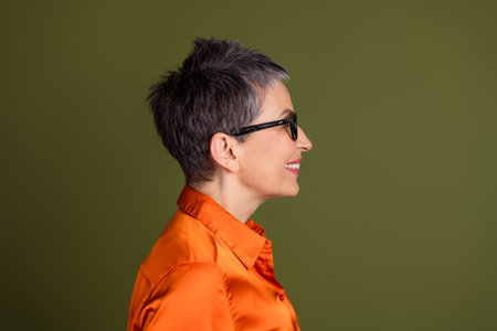 Profile of a mature woman with short gray hair wearing glasses and an orange shirt against a plain green backgroundの写真素材