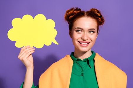 Stylish red haired woman in green outfit holding blank yellow speech bubble against vibrant purple...