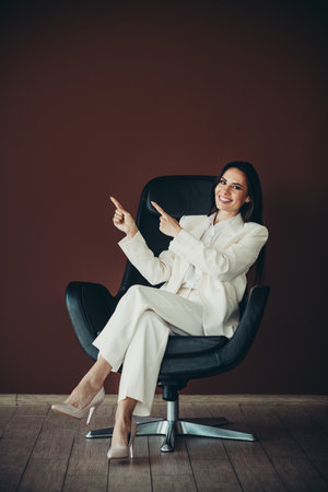 Confident young businesswoman in a stylish white suit sitting on an office chair with a welcoming smile and gestureの写真素材