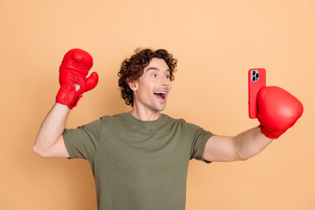 Young man with curly hair in casual style posing with boxing gloves and smartphone in front of a beige backgroundの写真素材