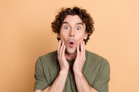 Young man with surprised expression wearing green t-shirt posing against beige background in a playful casual portraitの写真素材