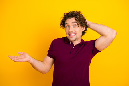 Confident young man with curly hair wearing burgundy polo gestures with surprise against a bright yellow backgroundの写真素材