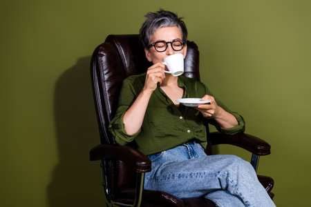 Mature woman enjoying leisure time with a cup of coffee wearing stylish casual outfit while sitting comfortably in an armchairの写真素材
