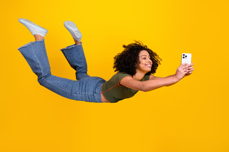 Young woman on yellow background posing playfully with a smartphone, capturing joyful moments in a...