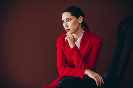 Attractive Confident Businesswoman in Red Suit Posing on Chair Against Brown Backgroundの写真素材