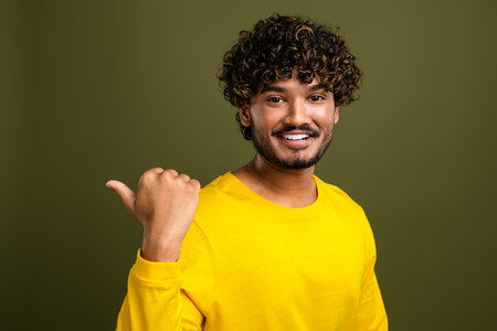 Cheerful young man gestures thumbs up in bright yellow shirt with curly hair and beard against green backgroundの写真素材