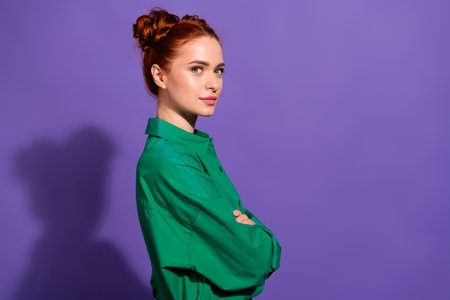 Confident young woman in casual green shirt posing on vibrant purple background with stylish red hair bunの写真素材