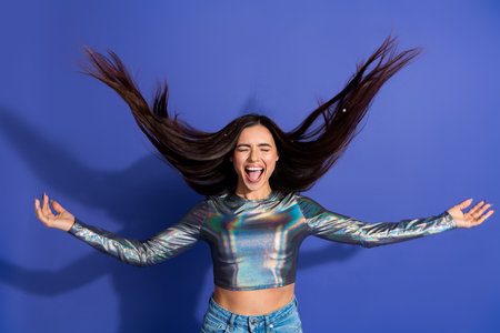 Cheerful young woman with glossy top celebrating against vibrant purple background displaying dynamic hair movement and joyの写真素材