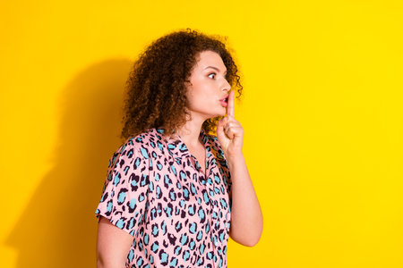 Young stylish woman gestures for silence with curly hair and leopard print shirt against a bright...