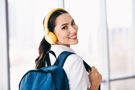 Charming woman with a backpack and headphones smiling confidently in a bright modern urban settingの写真素材