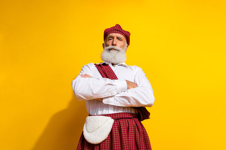 Confident bearded man in Scottish attire posing against yellow background with arms crossed, showcasing traditional styleの写真素材