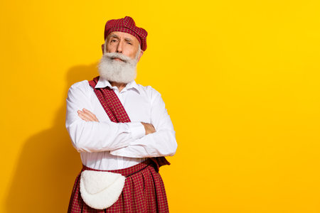 Scottish elder in traditional tartan outfit against vibrant yellow backdrop, expressing culture and confidence with folded arms.の写真素材