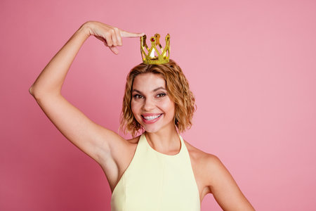 Smiling young woman in yellow dress wearing a fancy crown and posing against a pink background, exuding happinessの写真素材