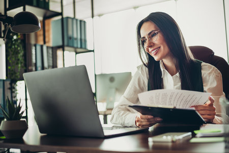 Confident businesswoman working in an office holding documents and smiling while sitting in professional workspace settingの写真素材