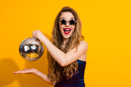 Excited young woman in star sunglasses holding a disco ball against a vibrant yellow backgroundの写真素材