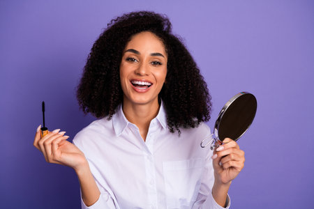 Charming young woman holding a mascara wand and a mirror on a purple background, showcasing beauty...