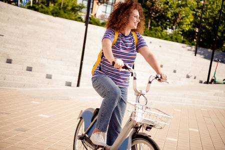 Young woman with curly hair riding a bike in the city, enjoying a bright summer day, wearing a...
