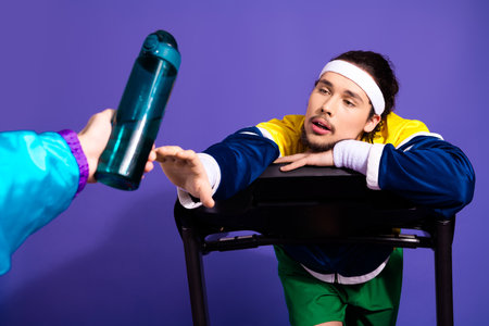 Retro Styled Man in Activewear Reaching for Water Bottle on Purple Backdrop in Sporty Training...