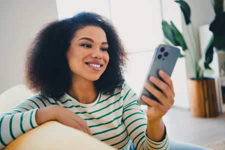 Young woman with curly hair smiling at her smartphone while relaxing on a cozy couch in a bright modern living roomの写真素材