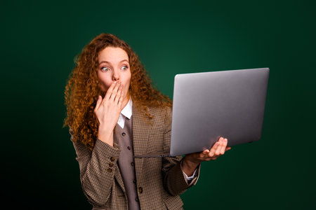 Surprised businesswoman with a laptop in formal attire against a green backdrop gesturing with hand to cover a shockの写真素材