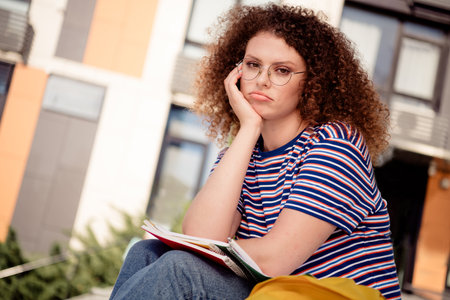 Young student with curly hair sitting outdoors, wearing striped t-shirt, looking thoughtful on a...