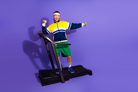 Young man in retro sportswear exercising on a treadmill with dumbbells on a vibrant purple...